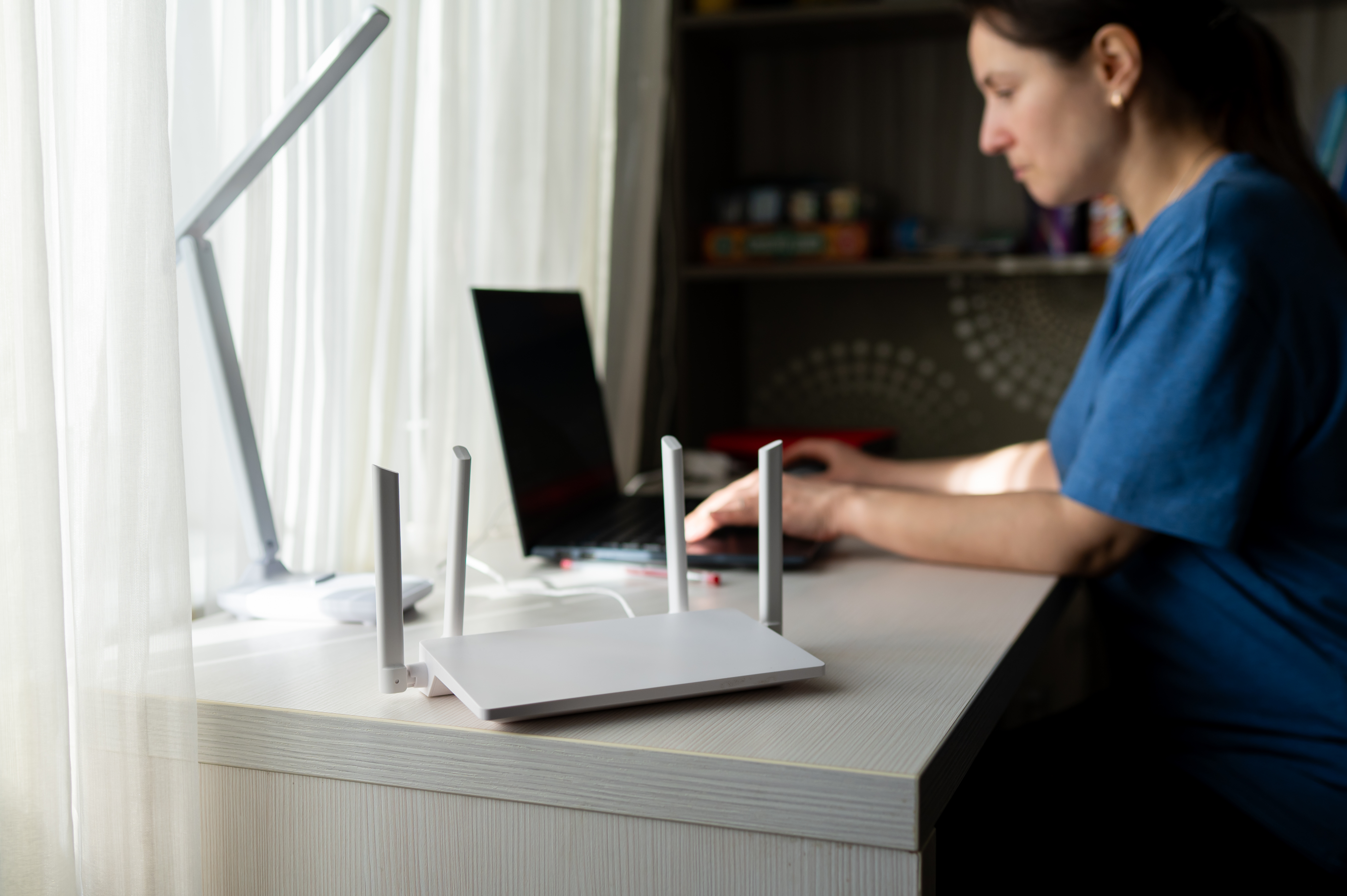 Close-up of a modern wireless router with antennas on a wooden desk. In the blurred background, a young woman is working on a laptop, representing high-speed internet and remote work lifestyle