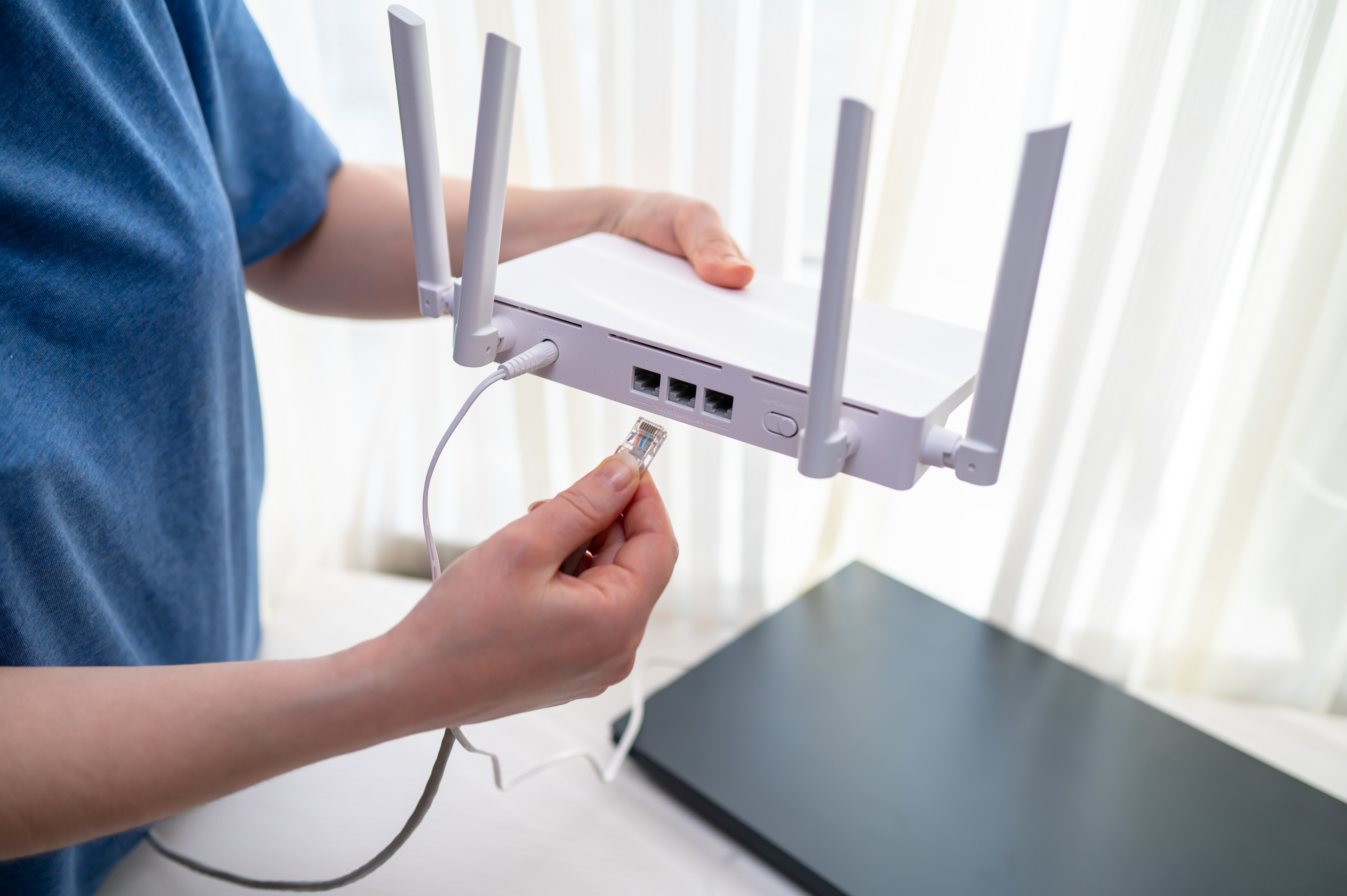 Close-up of a person in a blue shirt connecting a network cable into a wireless router with antennas. Concept of high speed internet setup, remote work workspace and home network installation